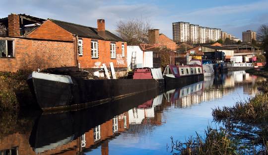 Canal-side view with moored narrowboats, red-brick warehouses, and reflections of the buildings on calm water.