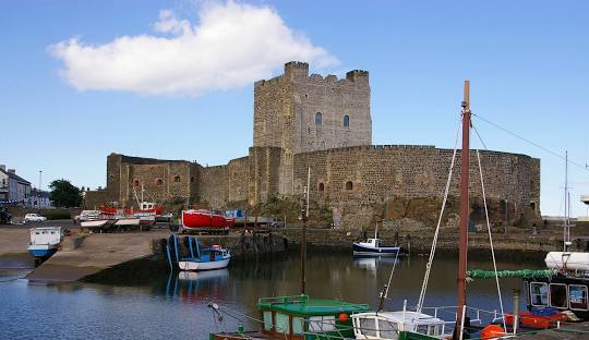 Historic stone castle beside a small harbor with colorful fishing boats moored in calm waters under a bright blue sky.