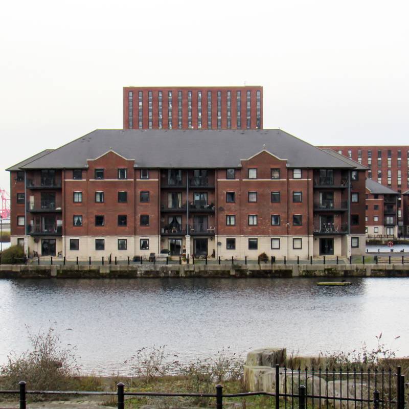 Waterside residential complex featuring red brick apartment buildings with balconies overlooking a calm river on an overcast day.