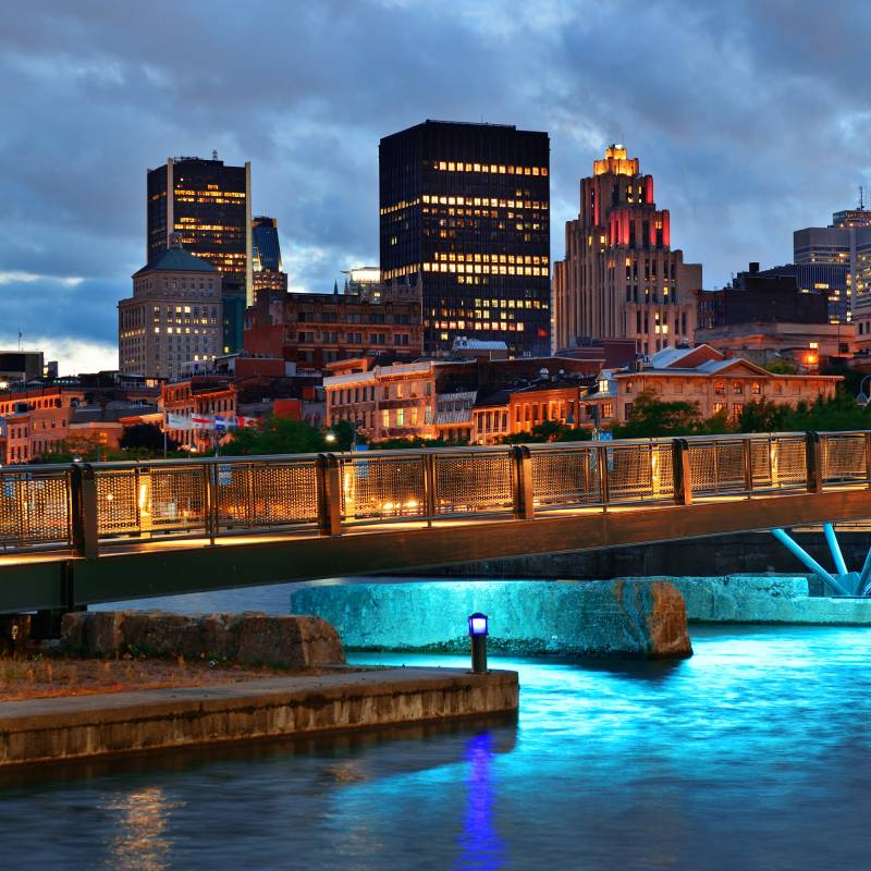 Illuminated urban skyline at dusk with tall modern buildings, a lit pedestrian bridge, and reflections on the water below.