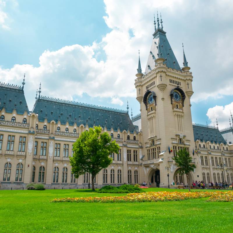 Grand historic building with a tall clock tower, ornate architecture, manicured green lawns, and flowerbeds under a partly cloudy sky.