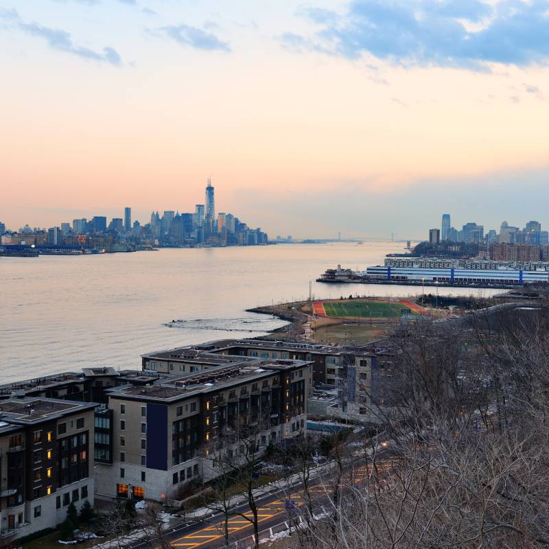 Scenic waterfront view at sunset featuring a city skyline in the distance, modern apartment buildings in the foreground.